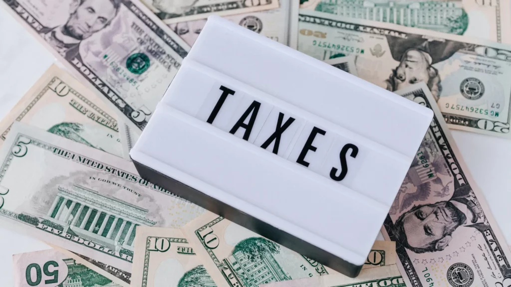 The image shows a lightbox with the word "TAXES" in bold letters placed on top of a spread of various denominations of U.S. dollar bills.