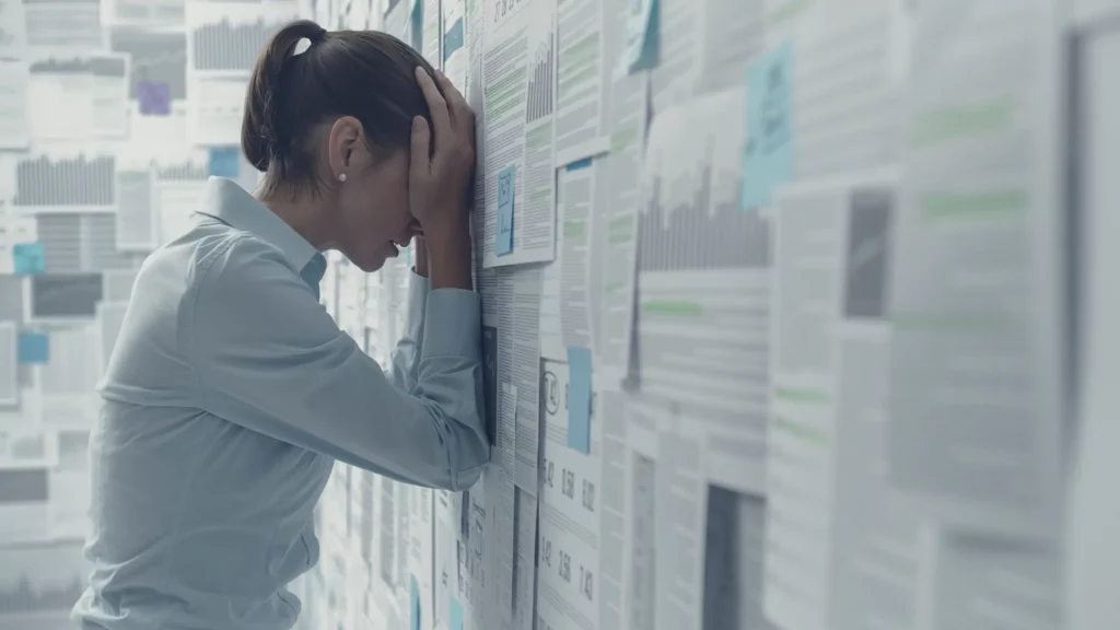 A woman in a light blue shirt is standing with her head against a wall covered in printed spreadsheets and graphs, holding her head in her hands in a gesture of frustration or exhaustion.