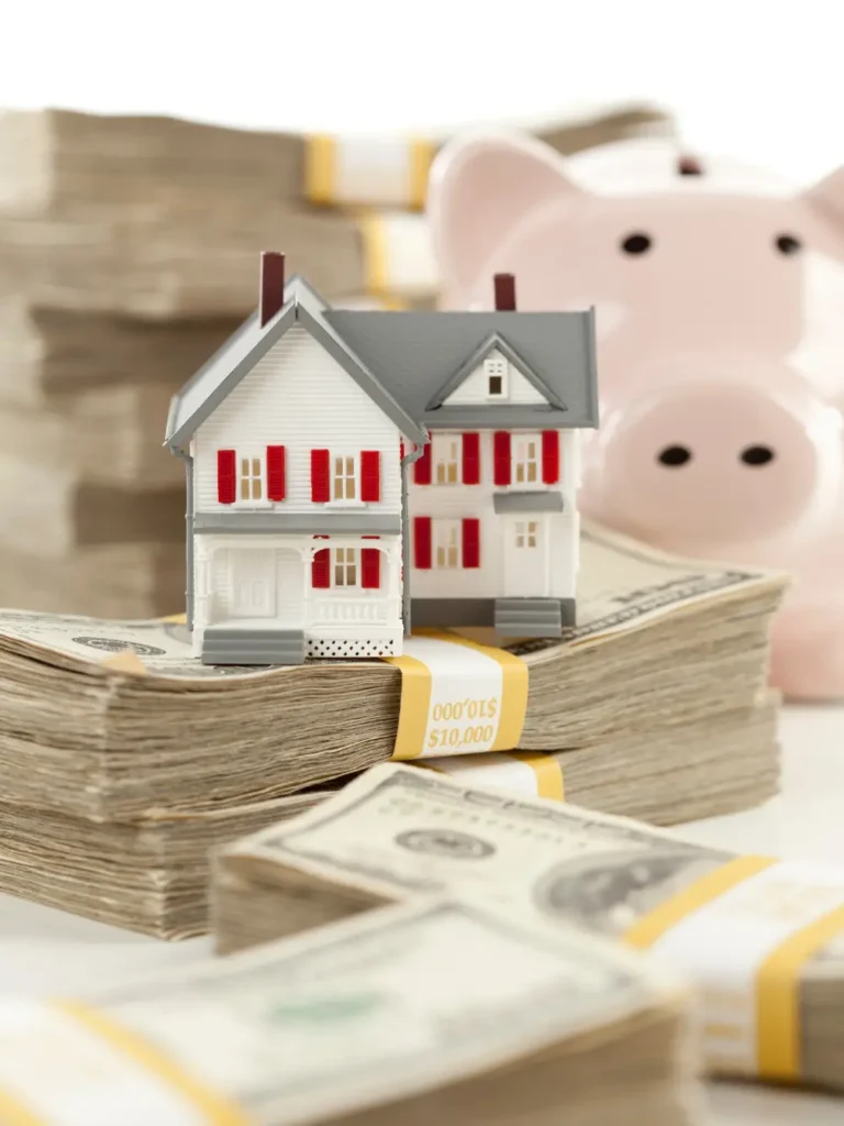 A miniature model of a two-story house with red shutters is placed on top of neatly stacked bundles of US hundred-dollar bills. In the blurred background, there is a pink piggy bank.