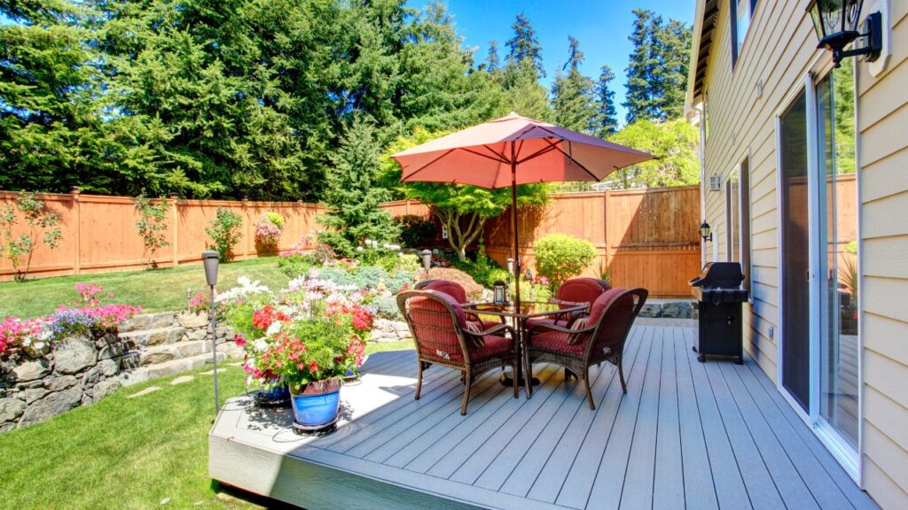 A sunny backyard patio with a dining set comprising a table and four wicker chairs under a large red umbrella. The patio is bordered by a vibrant garden full of pink and red flowers and a lush green lawn, all enclosed by a tall wooden fence. Tall trees stand in the background, and a barbecue grill is situated to the right, next to the siding of a house with sliding glass doors.