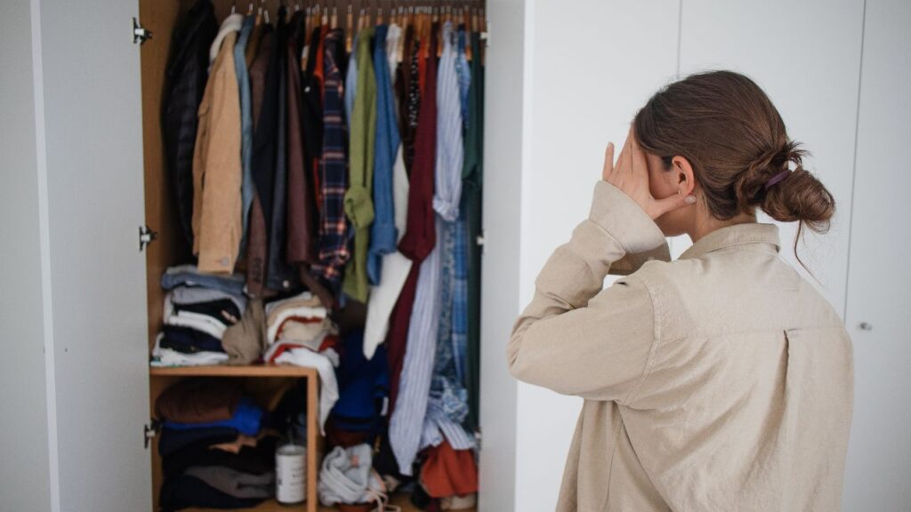 A woman stands in front of an open wardrobe, appearing overwhelmed by the cluttered and disorganized clothing items inside. The wardrobe is filled to the brim with clothes hanging on racks and stacked on shelves.