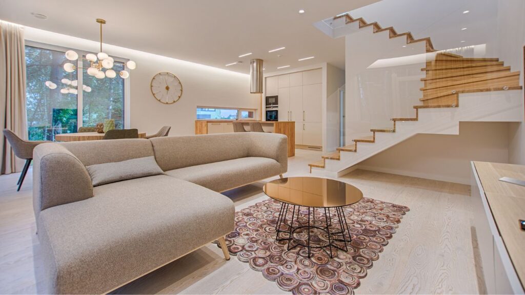 A modern living room interior featuring a beige sectional sofa, a round coffee table with a reflective surface on a decorative rug, and a wooden staircase with glass balustrades. The room has large windows with a view of trees, a sleek kitchen in the background, and a contemporary chandelier overhead.