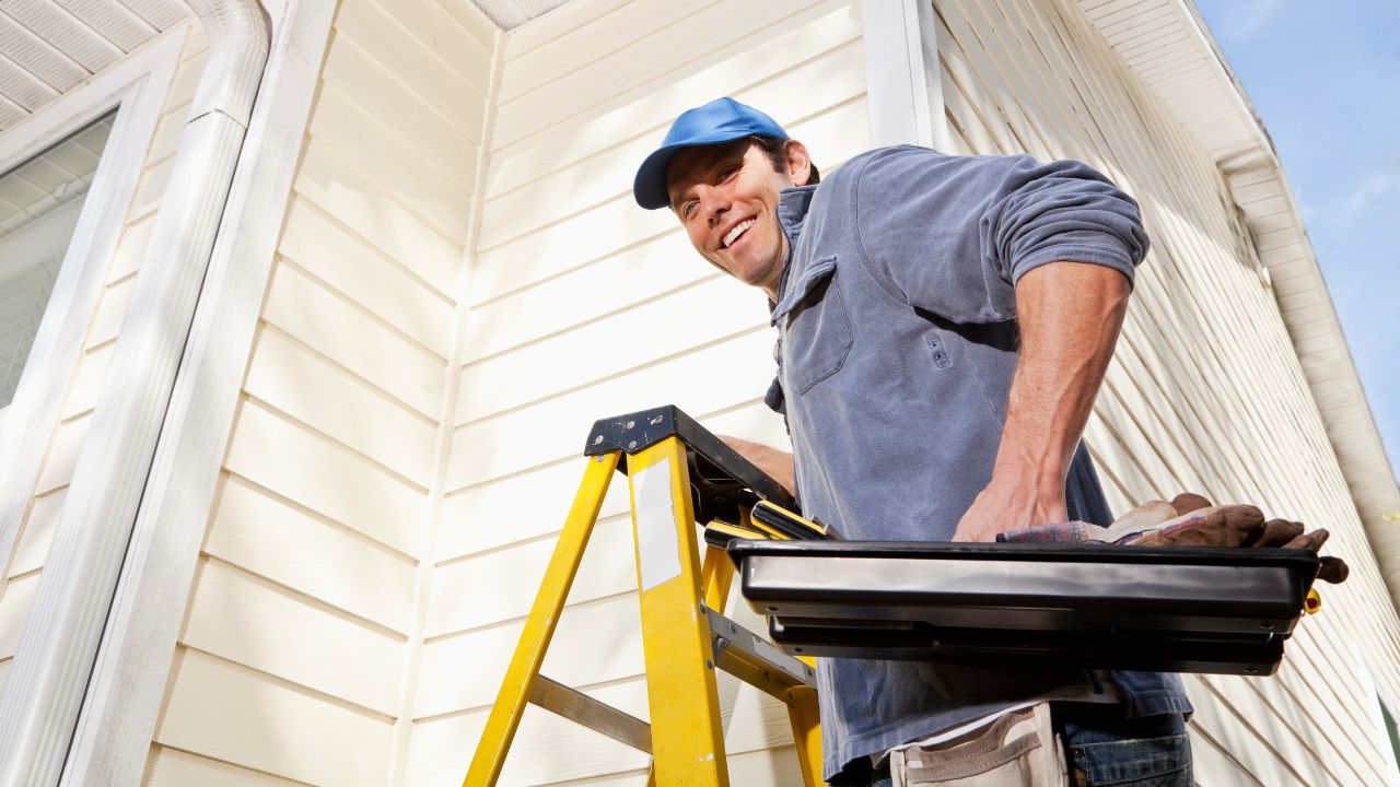 A smiling man wearing a blue cap and gray shirt is standing on a yellow stepladder outside a house with white siding. He is holding a tool tray filled with various items, suggesting he is engaged in home repair or maintenance work.
