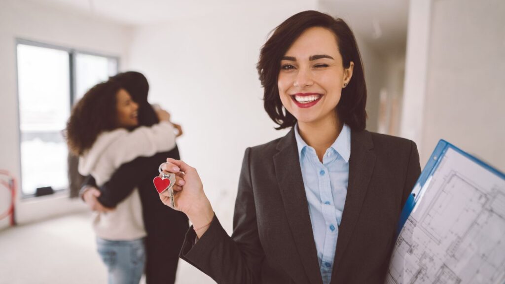 A smiling real estate agent holds up keys with a heart-shaped keychain, signifying a new home purchase. She is dressed professionally in a suit and holds blueprints or documents under her arm, indicating preparedness and competence in her role. In the background, a happy couple embraces, symbolizing their joy possibly at buying a new home, which is likely facilitated by the agent in the foreground. The focus on the keys and the agent’s smile suggests a successful real estate transaction.