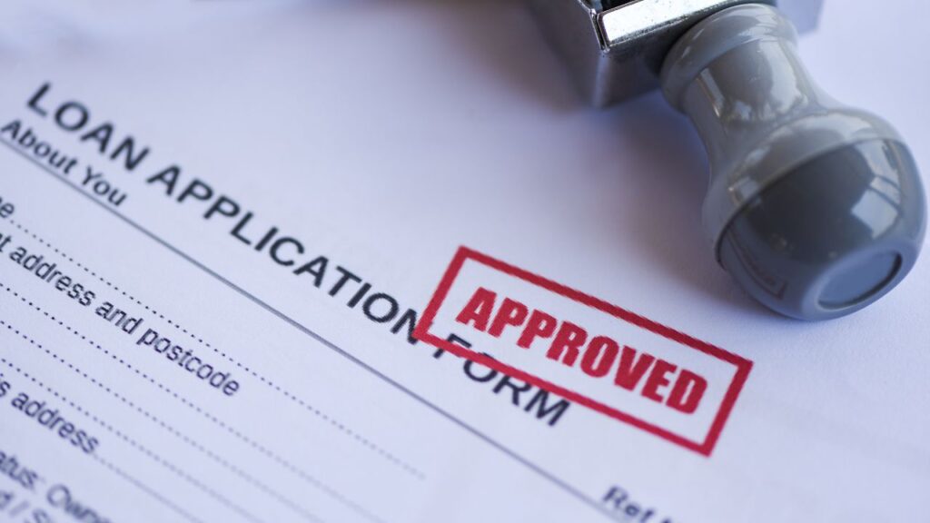 A loan application form with a section titled "About you" including fields for the applicant's address and postcode. The form has a red "APPROVED" stamp on it, indicating that the loan application has been accepted. There is also a blue and silver rubber stamp resting on the form, its handle visible and its stamping end resting on the paper.
