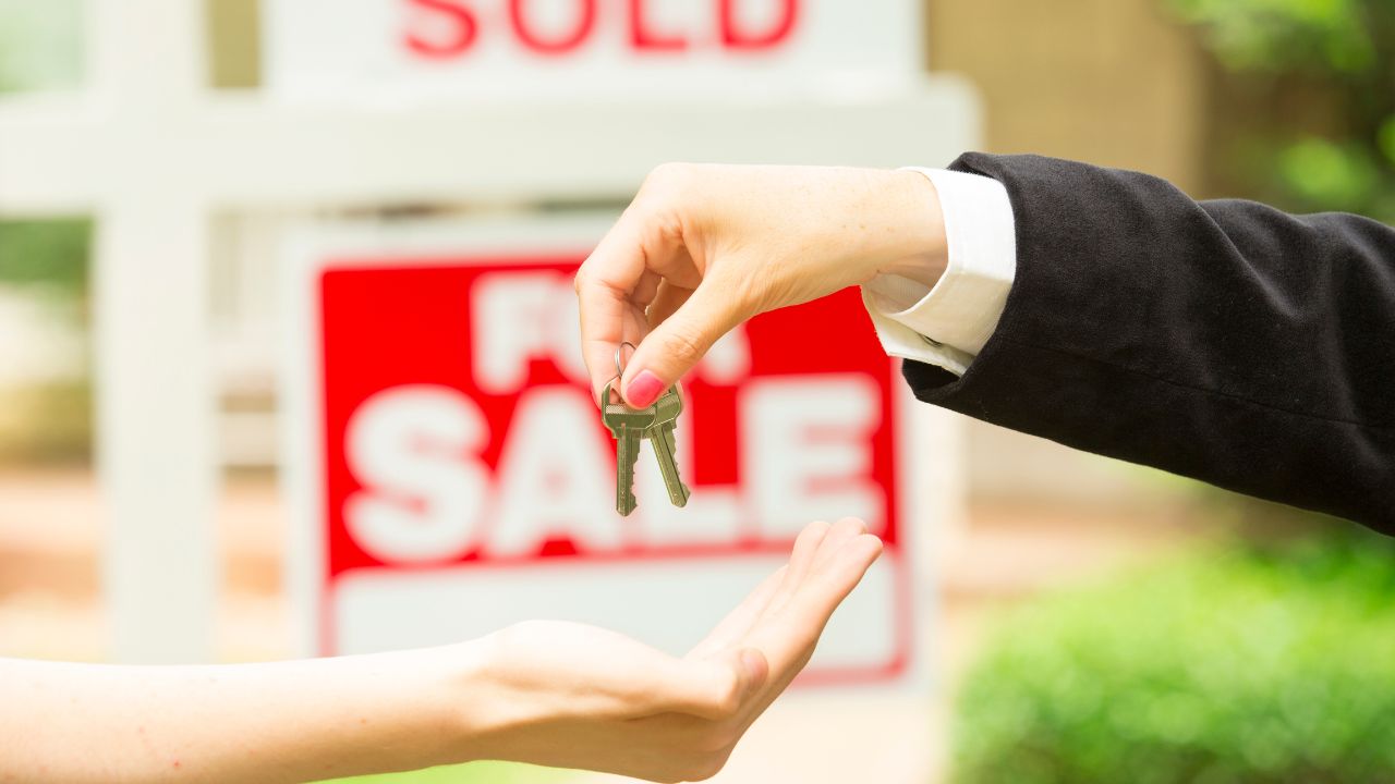 A close-up of two hands exchanging keys, with a 'SOLD' real estate sign in the background indicating a completed property sale.