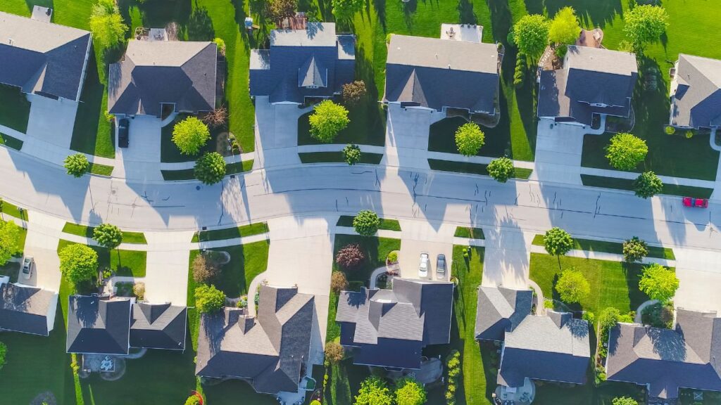 An aerial view of a suburban neighborhood with neatly arranged houses. The homes are situated along curved, tree-lined streets with well-maintained lawns and driveways. Each house has a similar architectural style and layout, showcasing a cohesive and organized community. The vibrant green lawns and scattered trees add to the picturesque and orderly appearance of the area.