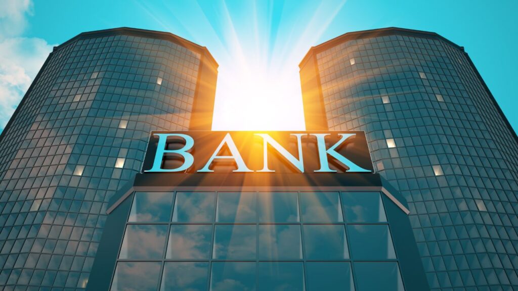 The image features a modern bank building with a reflective glass facade, consisting of two tall, cylindrical towers. The word "BANK" is prominently displayed on a sign at the entrance, highlighted by the bright sunlight shining from behind the building.