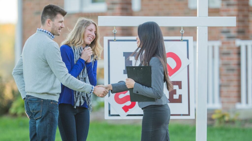 A happy couple is shaking hands with a real estate agent in front of a "For Sale" sign. The man is wearing a light gray sweater and jeans, while the woman is dressed in a blue sweater and scarf. The real estate agent is holding a clipboard and is dressed in a professional gray blazer and pants. They are standing outside in front of a house, suggesting they have just completed a successful property transaction.