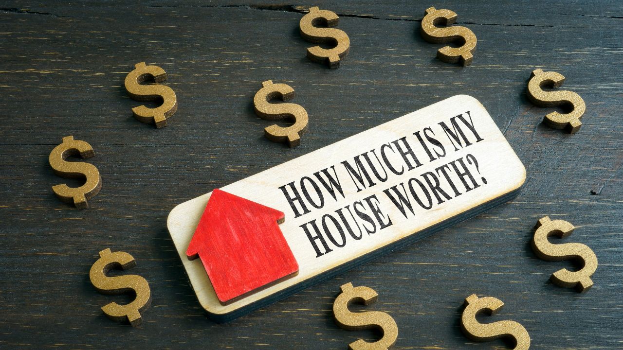 A wooden plaque with the text "HOW MUCH IS MY HOUSE WORTH?" is displayed on a dark wooden surface. The plaque features a red house icon on the left side. Surrounding the plaque are several gold-colored dollar sign symbols, emphasizing the theme of real estate value and financial assessment.