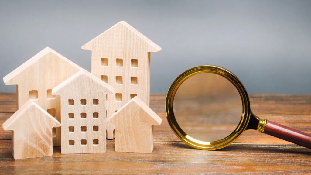 Wooden model houses of varying sizes are arranged on a wooden surface, with a large magnifying glass positioned to the right of the houses. The magnifying glass has a golden frame and a wooden handle, suggesting the concept of close inspection or valuation of the properties. The background is neutral, keeping the focus on the houses and the magnifying glass, symbolizing the process of assessing real estate value.