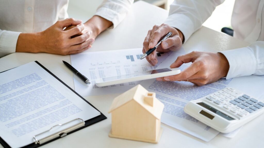 The image shows two people sitting at a table engaged in a discussion, likely related to real estate or financial planning. One person is holding a pen and pointing at a document with charts and numbers, possibly explaining or reviewing details. The other person is sitting with their hands clasped, listening attentively. On the table, there are various items including a clipboard with a document, a small wooden model of a house, a calculator, and more papers. The overall scene suggests a professional consultation, possibly regarding a real estate transaction, mortgage, or financial advice.