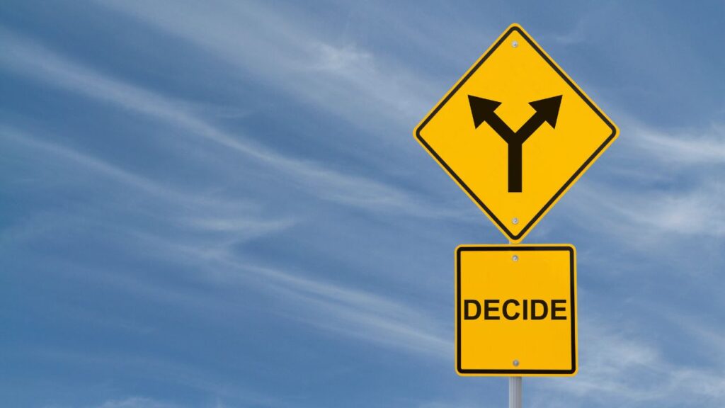 The image depicts two yellow road signs against a clear blue sky. The top sign is a diamond-shaped warning sign with a black symbol showing a fork in the road, indicating a split path or a decision point ahead. Below it is a rectangular sign with the word "DECIDE" in bold black letters. 