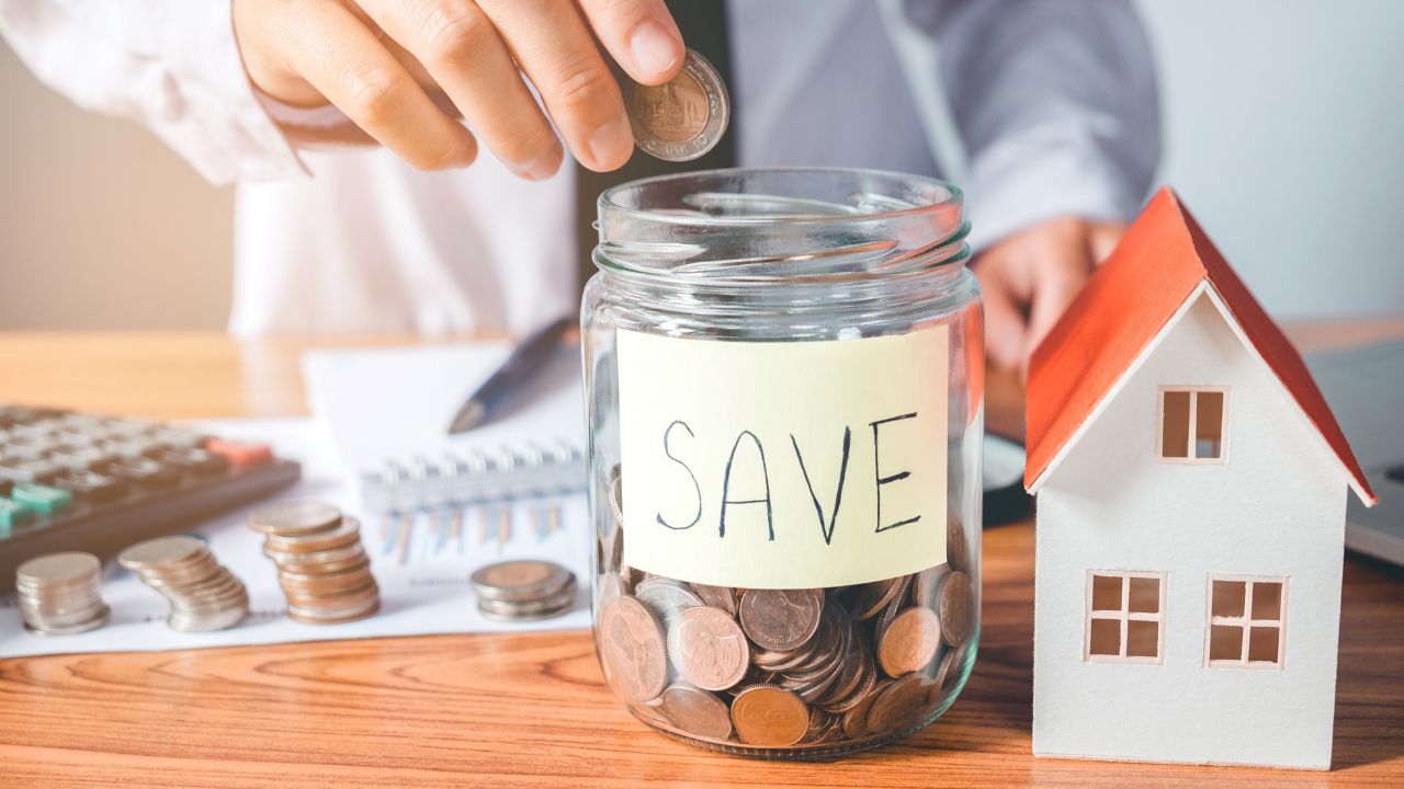 The image shows a person placing a coin into a clear glass jar labeled "SAVE." The jar is filled with coins, symbolizing savings. On the table, there are more coins stacked, a calculator, a pen, a notepad with charts, and a small model of a house, representing financial planning for homeownership.