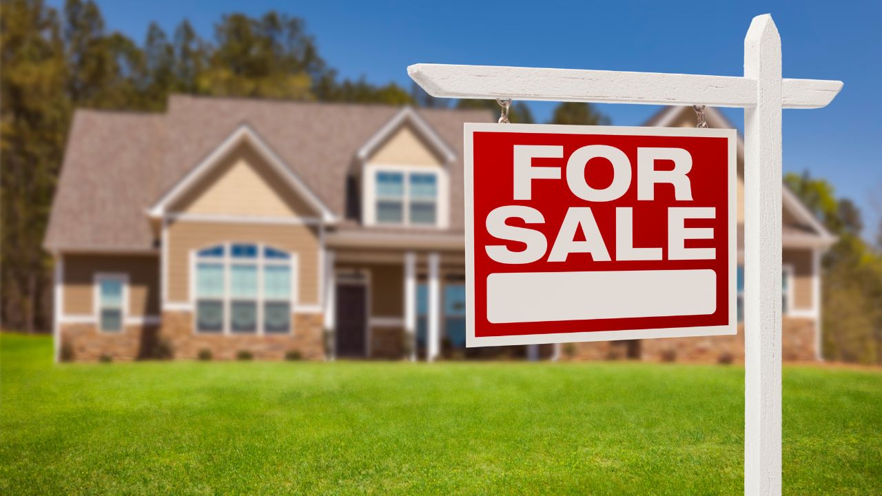 The image shows a large, white real estate sign with a red "FOR SALE" message in bold white letters. The sign is in the foreground, while in the background, a suburban house is slightly blurred. The house has a beige exterior with stone accents, large windows, and a well-maintained lawn.