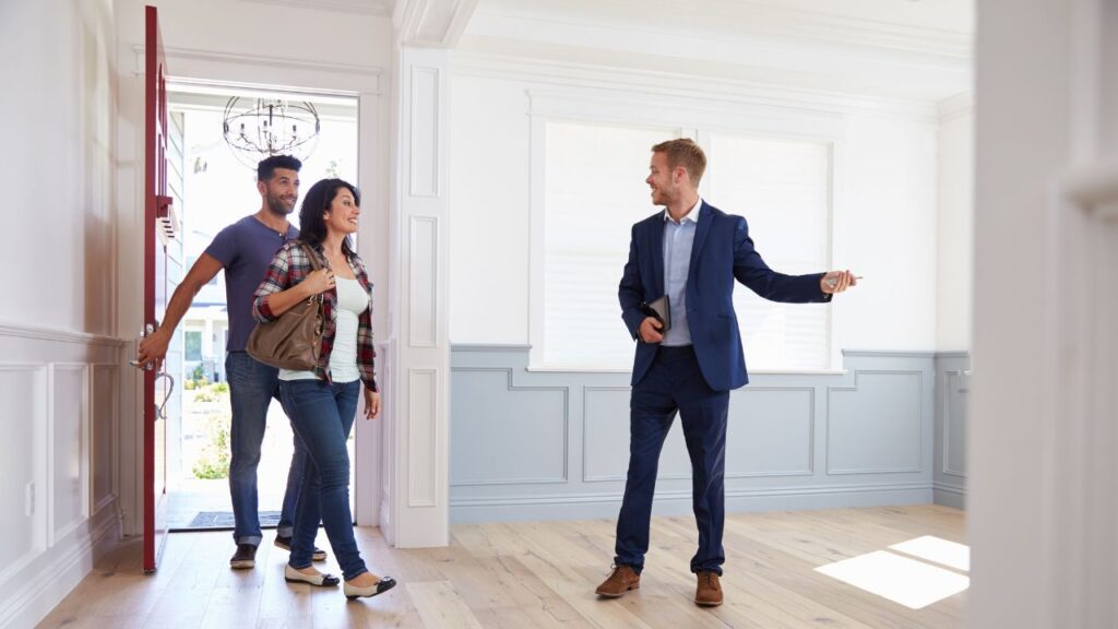 The image shows a real estate agent welcoming a couple into a bright, spacious room, likely during a home showing. The couple, a man and a woman, are entering through the front door with smiles on their faces. The man is holding the door open while the woman walks in first, carrying a handbag. The real estate agent, dressed in a navy blue suit, is standing inside the room, gesturing with one hand, possibly pointing out features of the home or inviting them further inside. The room has light-colored hardwood floors, white walls with wainscoting, and large windows that allow natural light to flood the space, creating a welcoming atmosphere.