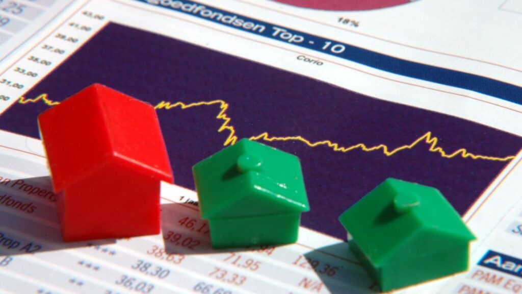 The image shows small red and green toy houses placed on top of a financial chart, with a fluctuating graph in the background.