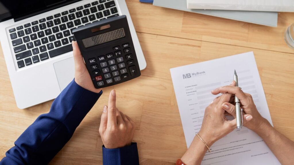 Close-up view of two people reviewing financial information at a desk. One person holds a large calculator displaying a number, while the other holds a pen, preparing to sign a document labeled 'MyBank' next to a laptop.