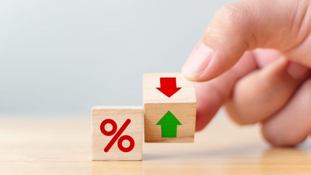 The image shows a close-up of a hand holding two wooden blocks. One block has a red percentage symbol, and the other has red and green arrows (down and up, respectively), representing the concept of changing interest rates or percentages.