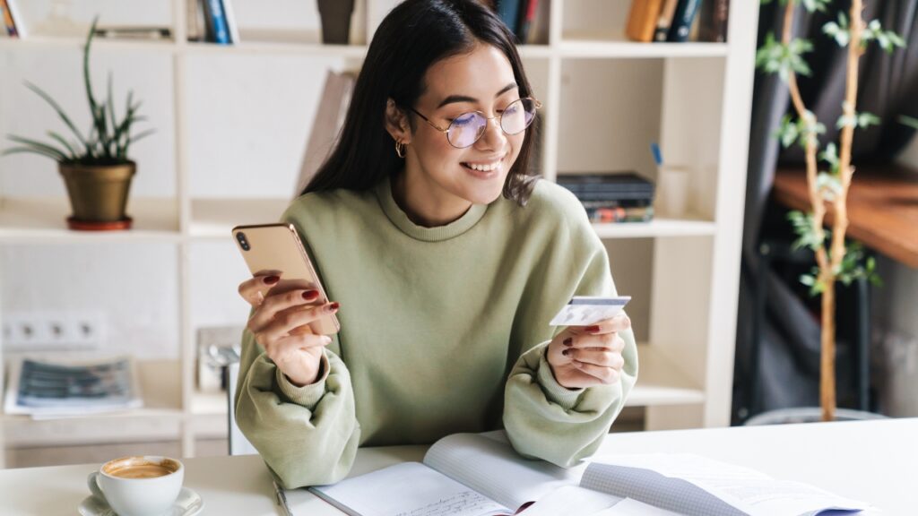 A young woman wearing glasses and a green sweater, sitting at a desk with an open notebook, holding a credit card in one hand and a smartphone in the other while smiling.