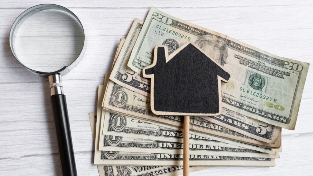 A magnifying glass and a black house-shaped cutout on top of a pile of U.S. dollar bills.