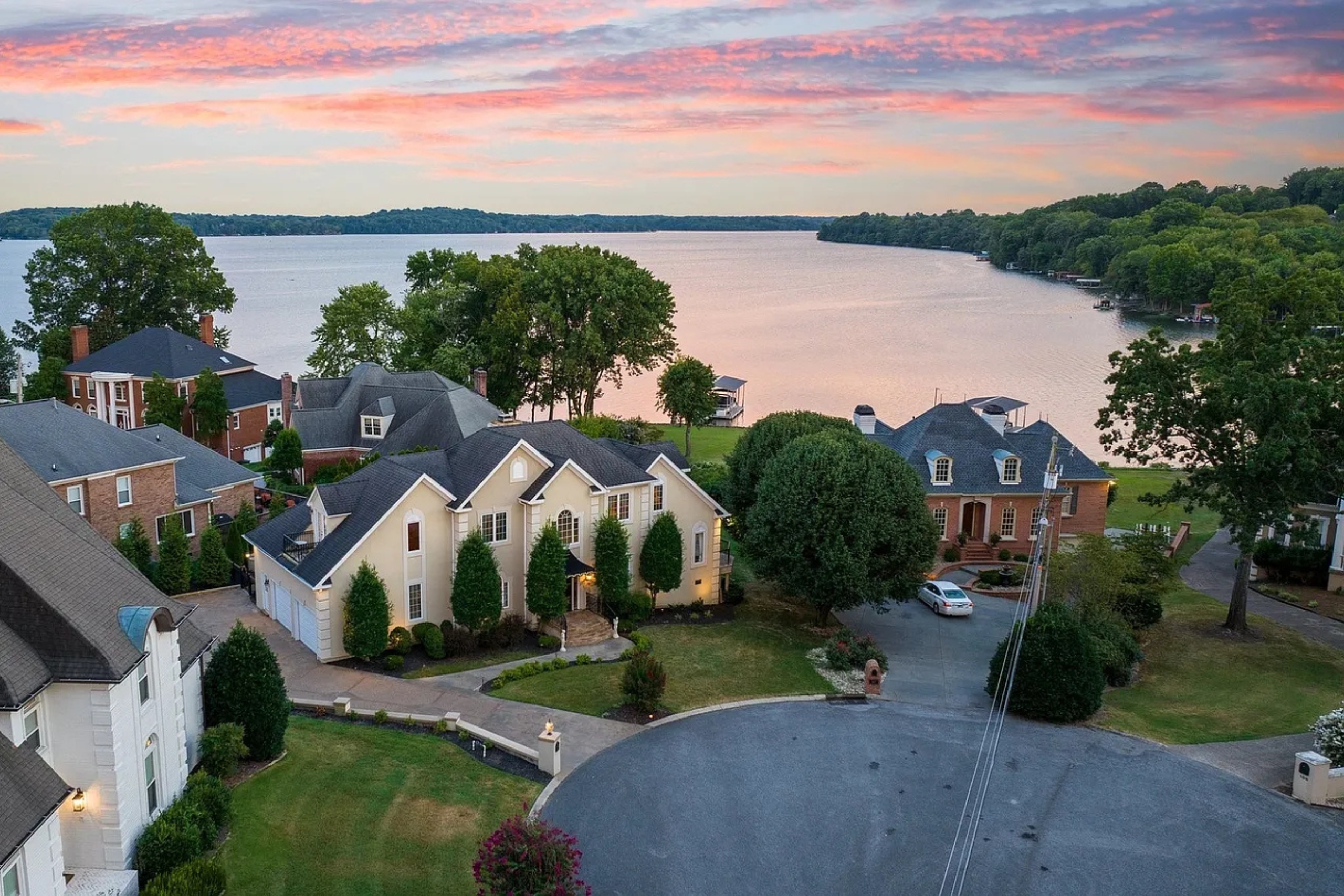 Aerial view of a quiet cul-de-sac lined with upscale homes along a serene lakefront in Hendersonville at sunset, with vibrant pink and orange clouds reflecting off the water.
