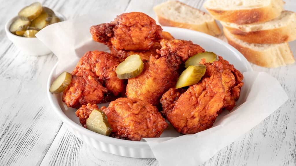A white bowl of Nashville hot chicken topped with pickles, served with sliced bread.
