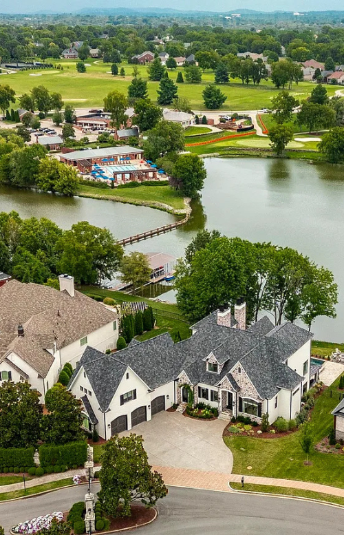 A large waterfront home sits at the end of a cul-de-sac with a golf course and clubhouse visible across the lake.