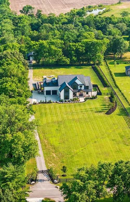 Aerial view of a modern farmhouse-style home on a large, well-manicured lot with fenced pastures and surrounding trees.
