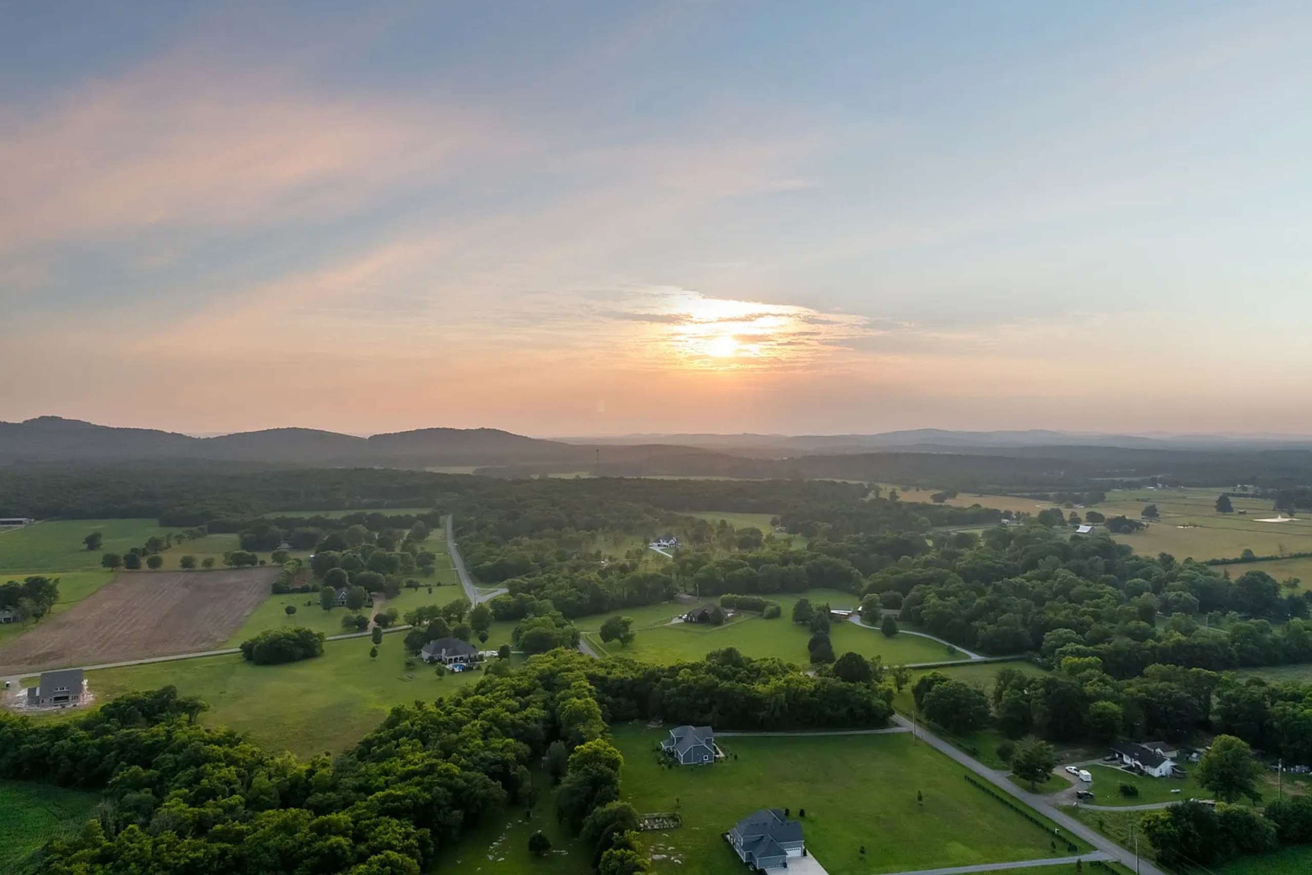 Aerial view of a rural landscape at sunrise with scattered houses, winding roads, and distant hills.