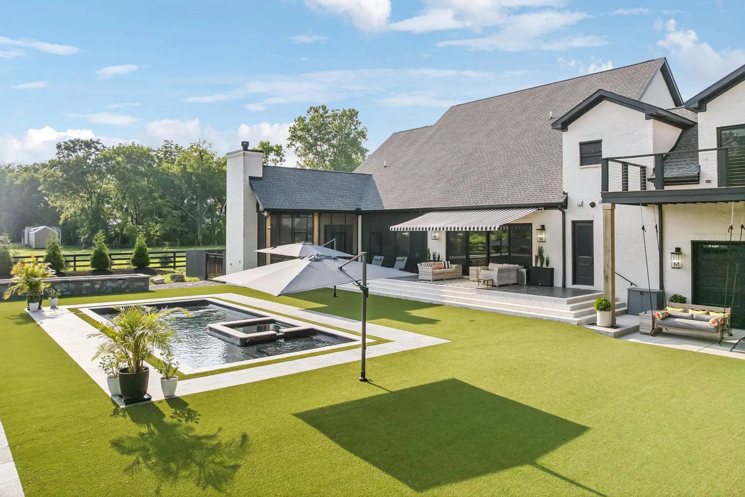 Backyard view of a modern white house with a small pool, patio seating, and artificial turf lawn.