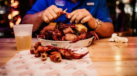 A man peeling crawfish at a table with a tray of crawfish, potatoes, and a cup of beer.