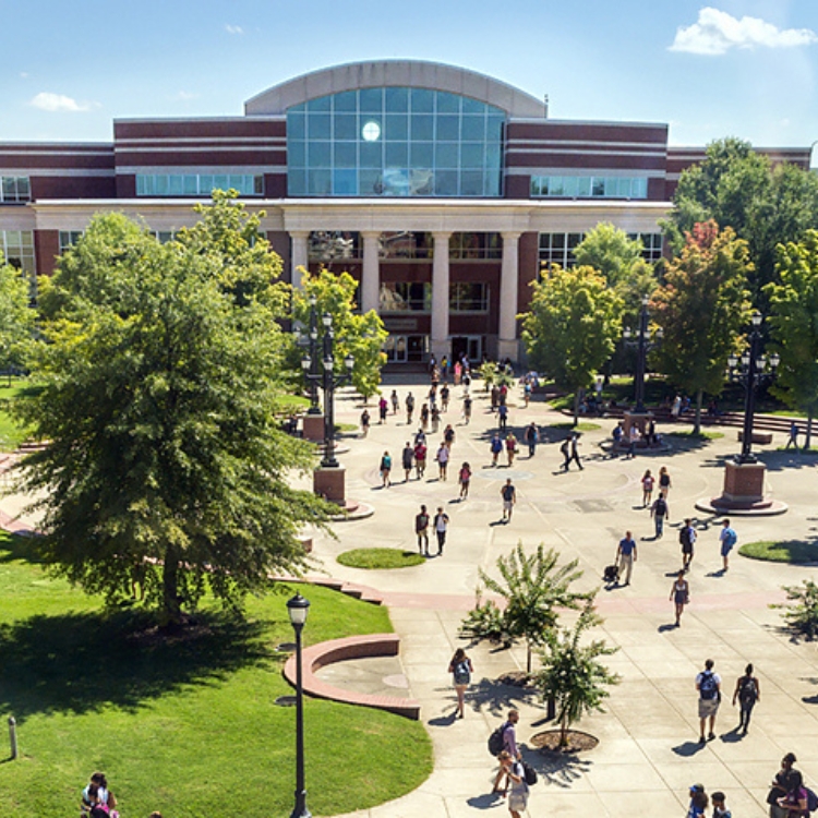 Students walk across a wide campus plaza toward a large modern academic building with tall glass windows and brick walls.
