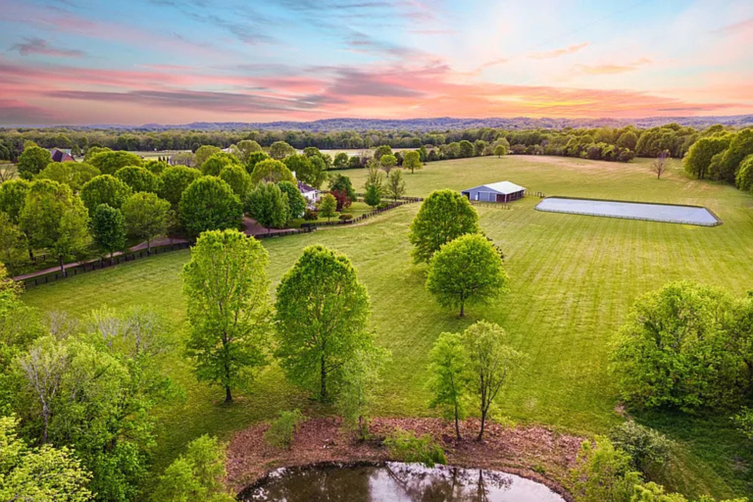 Aerial view of a large green property with a barn, pond, and trees at sunset.