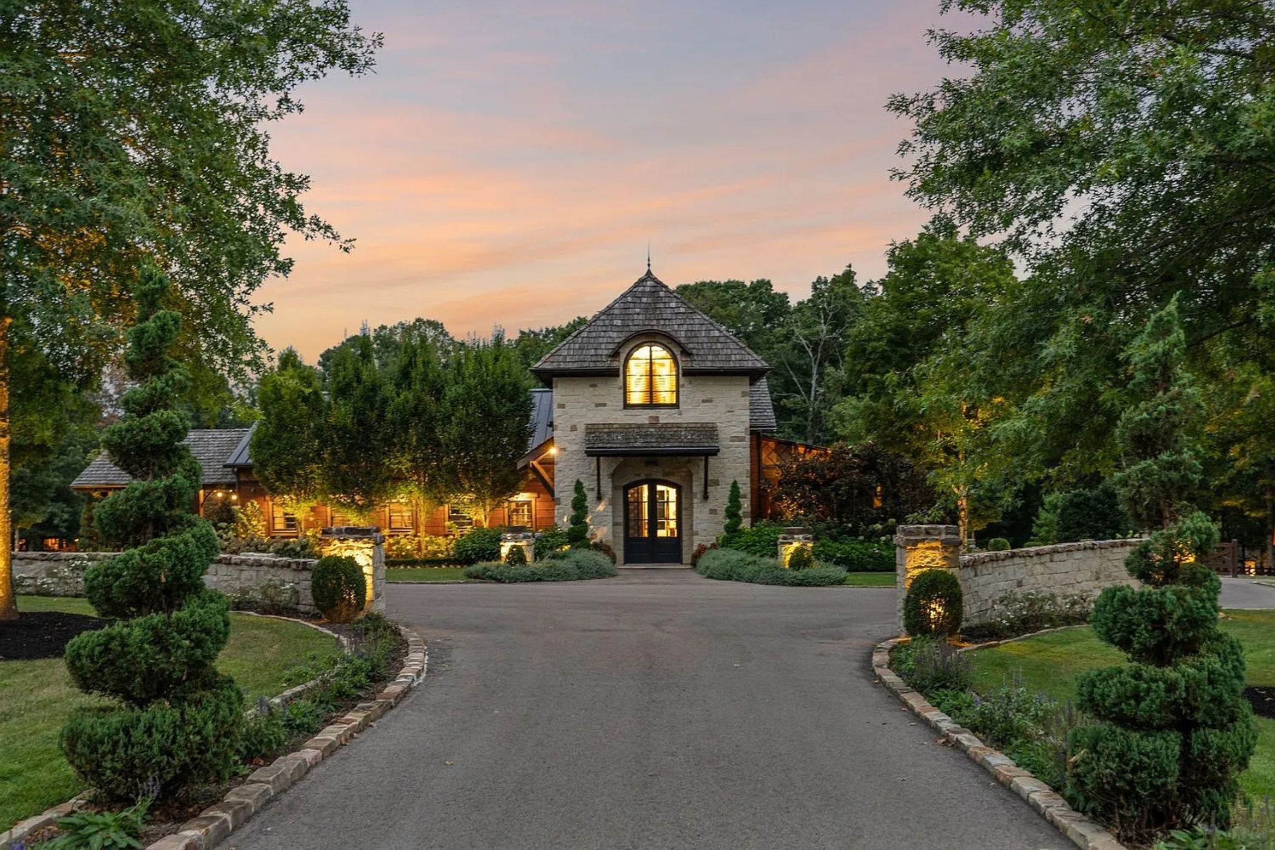 Elegant stone home with a turret-style entryway, lit landscaping, and circular driveway at dusk.