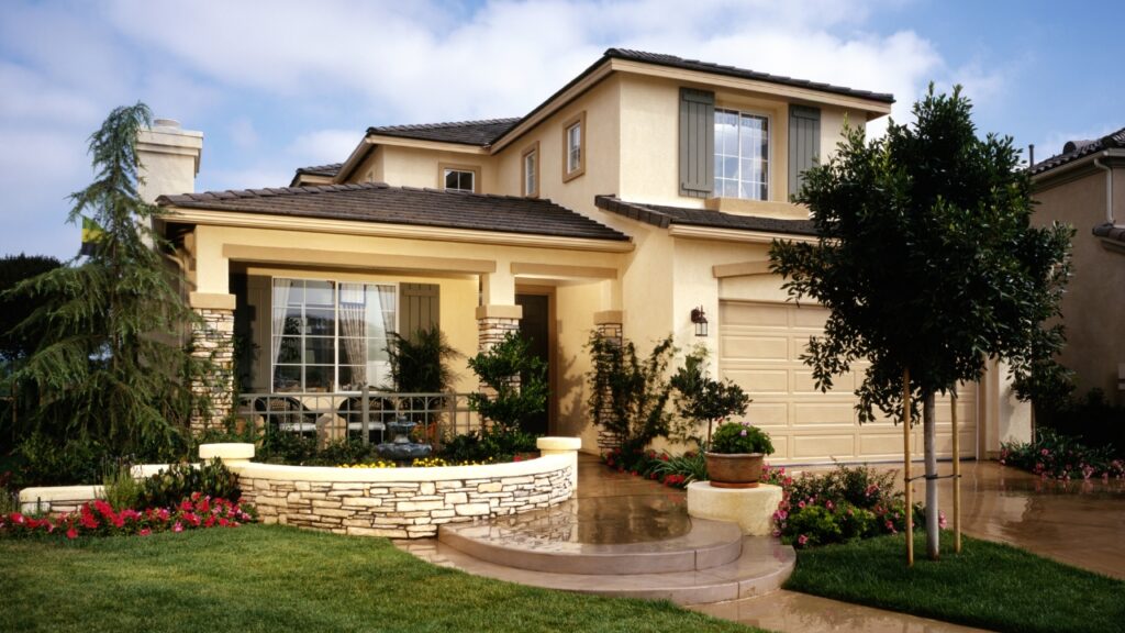 Beige two-story suburban house with landscaped front yard and large windows.