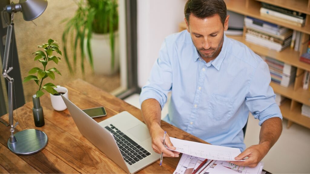 Man in a blue shirt reviewing documents at a desk with a laptop.