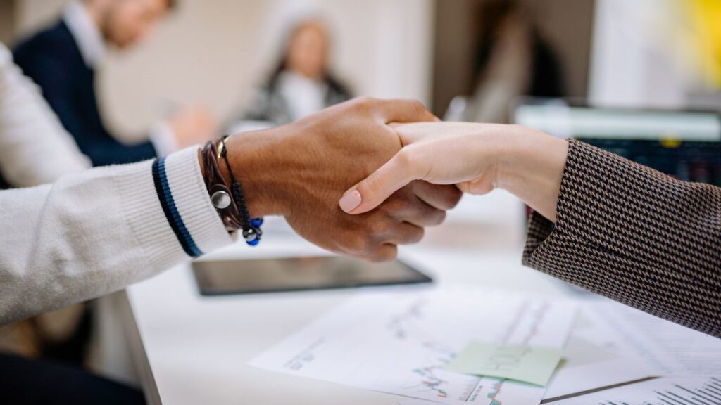 Two people shaking hands across a desk with papers and a tablet.