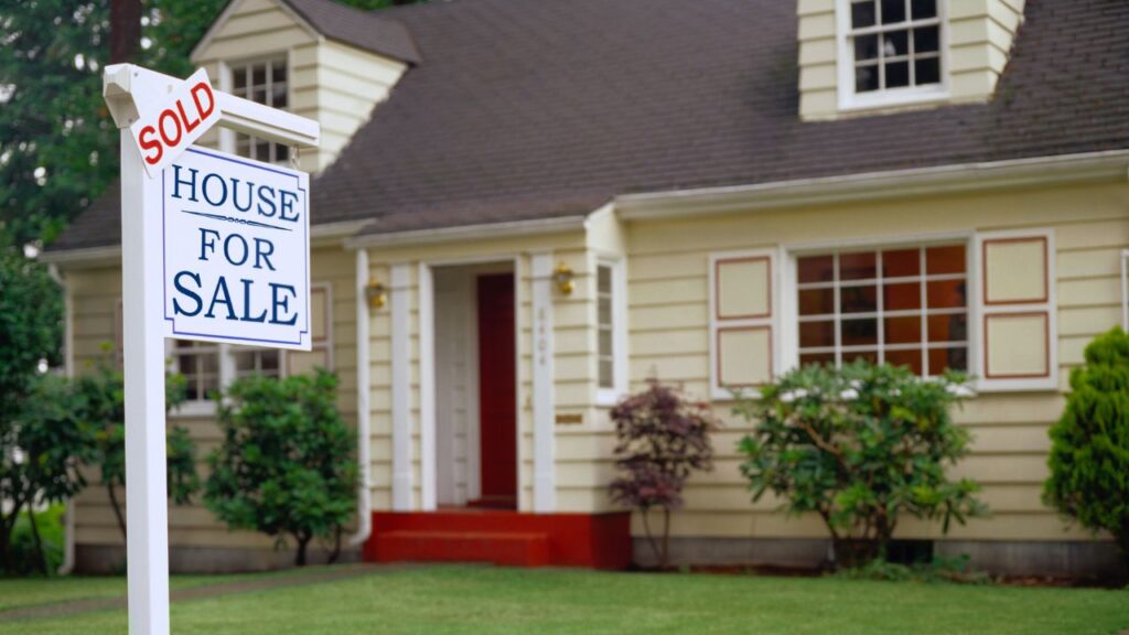 White house with a “House for Sale” sign that has a “Sold” label on top.