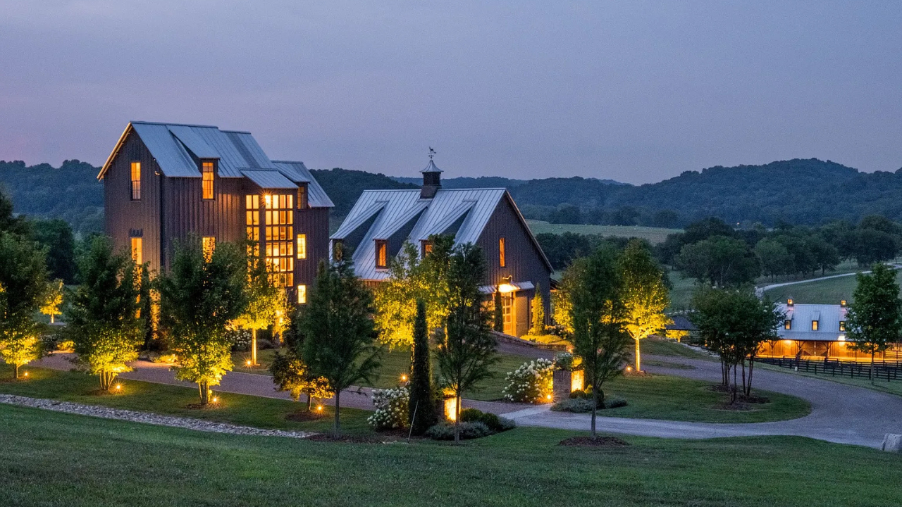 Large modern farmhouse estate with lit windows and landscaped driveway at dusk in rolling hills.