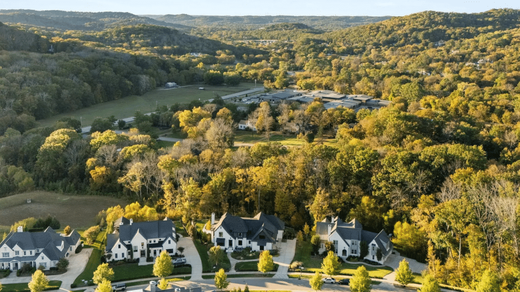 Aerial view of a upscale Brentwood, Tennessee neighborhood at golden hour in autumn, with large luxury homes nestled among tree-covered rolling hills and open green fields in the background.