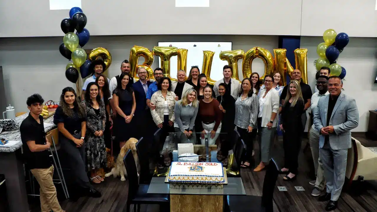 A large group of real estate professionals celebrating a $2 billion sold milestone, posing together with gold balloon letters and a decorated cake.